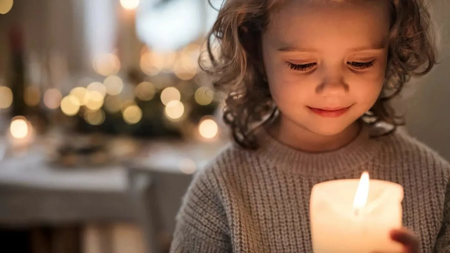 Une bougie blanche allumée, tenue à deux mains, illumine une pièce sombre avec des lumières floues en arrière-plan.