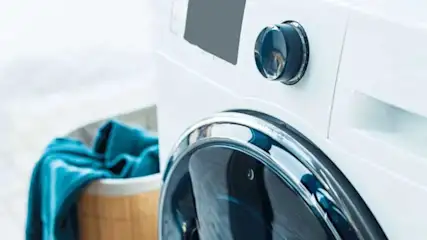 Een witte wasmachine met een chroomkleurige deur en een draaiknop. Naast de machine staat een houten wasmand met blauwe was.
