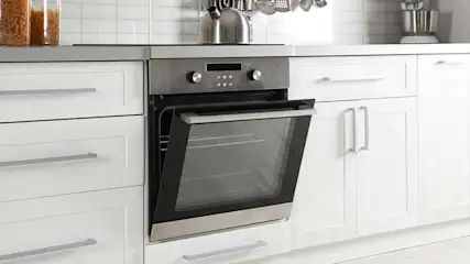 Een roestvrijstalen oven met open deur is ingebouwd in een witte keukenkast. Bovenop de kast staat een waterkoker.