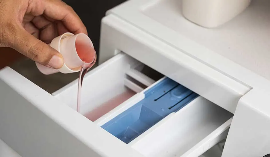 Hand giet roze vloeistof uit een maatbeker in een witte wasmachine dispenser met blauw compartiment.