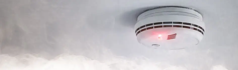Un détecteur de fumée blanc est fixé au plafond, avec une lumière rouge allumée et de la fumée visible.