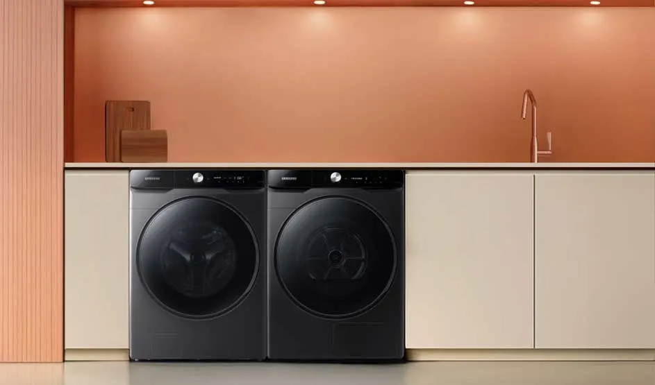 Minimalistische wasruimte: twee zwarte frontladers onder een wit werkblad, roze wand met spots, houten plankjes en een slanke kraan rechts.