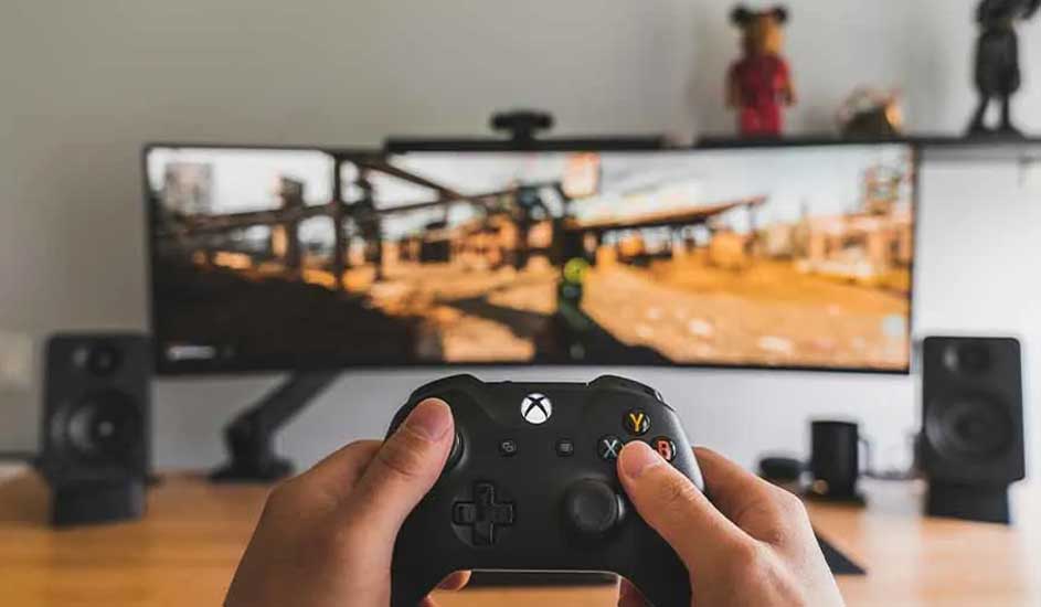 Deux mains tiennent une manette Xbox noire, pointée vers un écran large affichant un jeu vidéo, avec des haut-parleurs disposés de chaque côté.