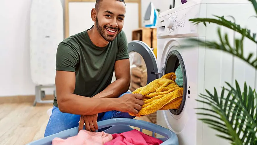 Un homme sort une serviette jaune d'un lave-linge blanc. Un panier à linge bleu est à côté de la machine.