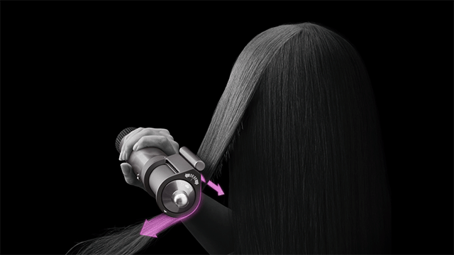 Sèche-cheveux gris utilisé sur des cheveux longs et lisses sur un fond noir, avec des flèches roses indiquant le mouvement.