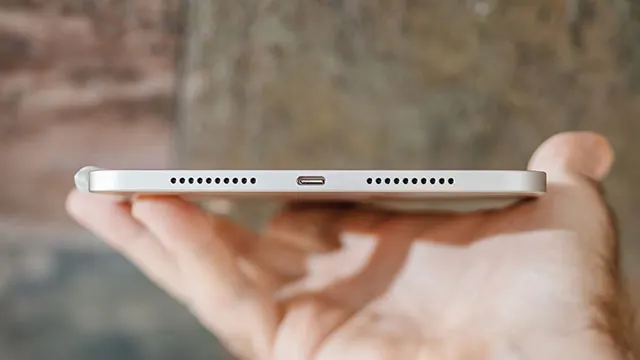 Un appareil électronique gris est tenu dans une main, avec un port de chargement et des grilles de haut-parleur visibles.