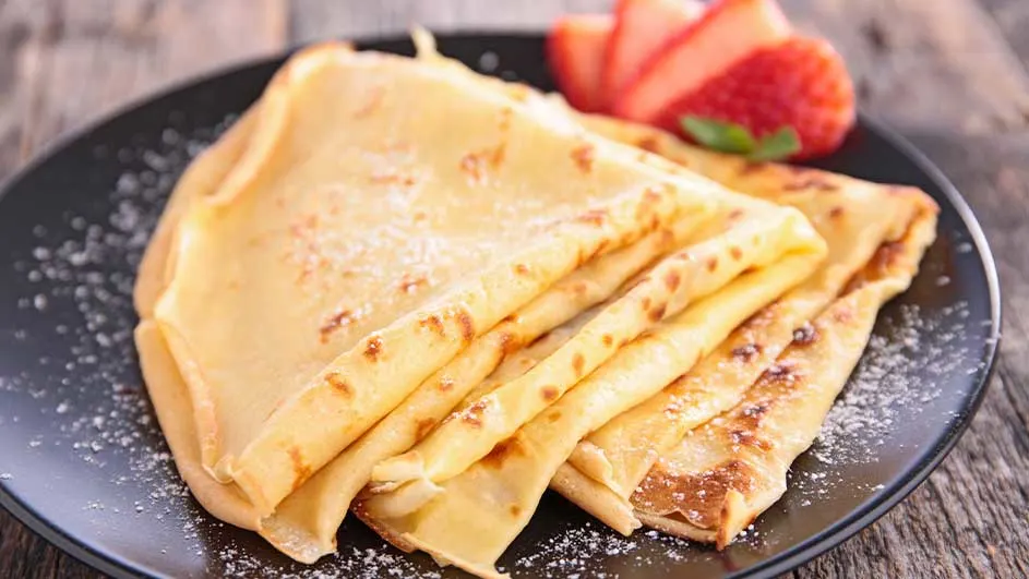 Stapel crêpes met aardbeien op een zwart bord, bestrooid met poedersuiker, op een houten tafel.