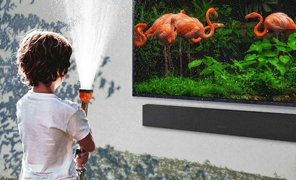 Jongen spuit met tuinslang voor tv met flamingo's. Zwarte soundbar hangt onder tv aan witte muur met blauwe schaduw.