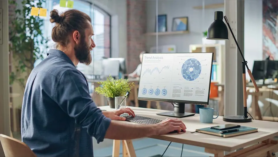 Een bureau met een computerscherm met grafieken, een lamp, een plant, een notitieboek en een blauwe mok.