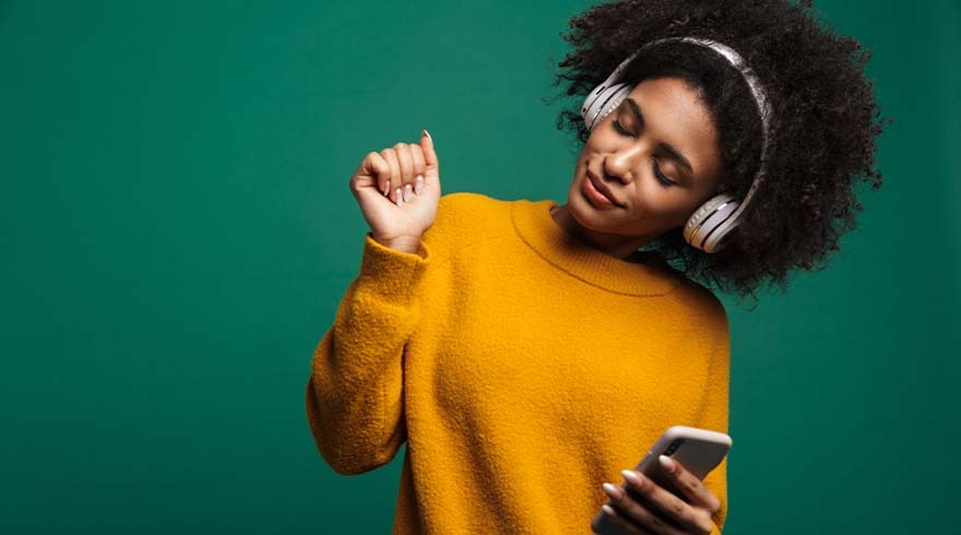 Une personne portant un pull jaune et un casque écoute de la musique sur un smartphone devant un fond vert.