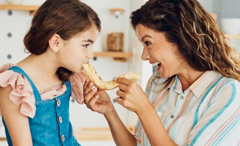 Une fille en salopette bleue mange une crêpe que lui donne une femme en chemise rayée. Arrière-plan clair.