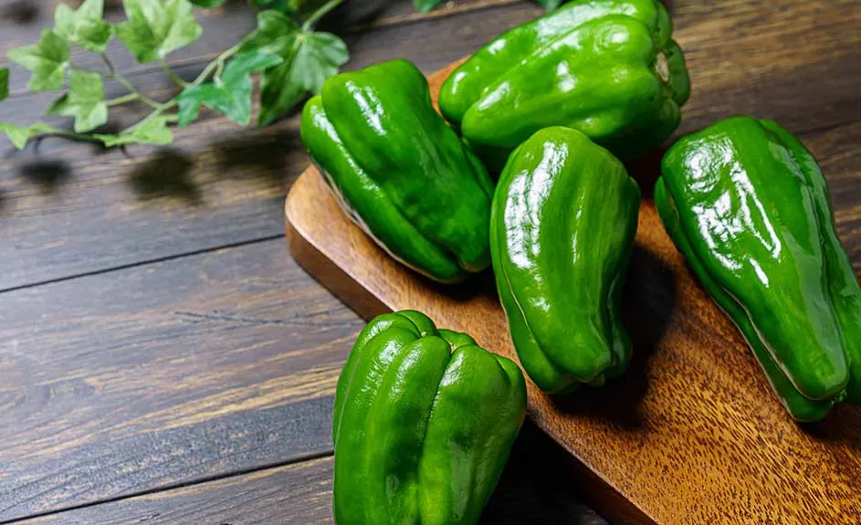 Cinq poivrons verts sur une planche à découper en bois brun, sur une table en bois avec une plante verte en arrière-plan.