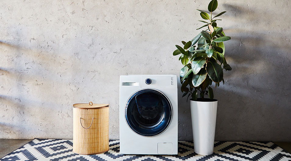 Un lave-linge blanc est à côté d'un panier à linge en osier et d'une plante en pot blanche sur un tapis à motifs géométriques.