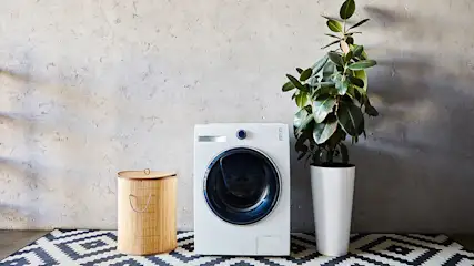 Un lave-linge blanc est à côté d'un panier à linge en osier et d'une plante en pot blanche sur un tapis à motifs géométriques.