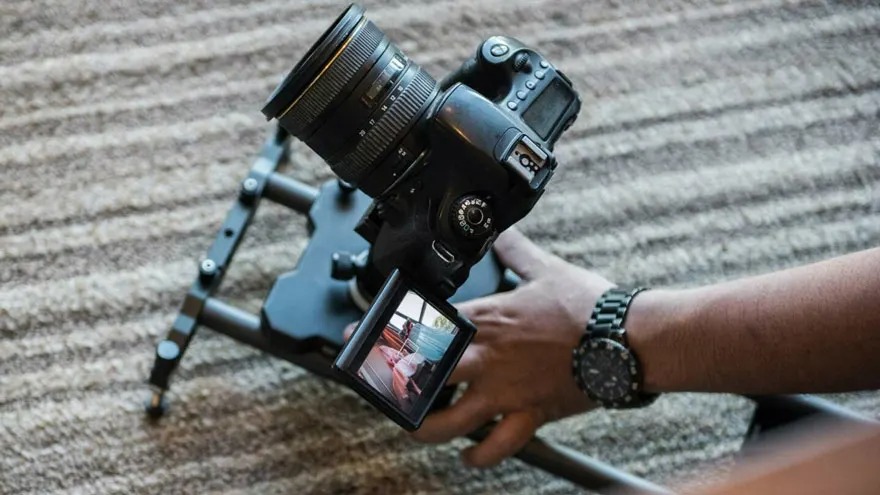 Een zwarte camera met lens op een statief, bediend door een hand met horloge, op een gestreepte achtergrond.