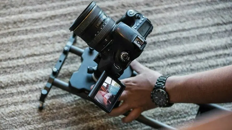 Een zwarte camera met lens op een statief, bediend door een hand met horloge, op een gestreepte achtergrond.