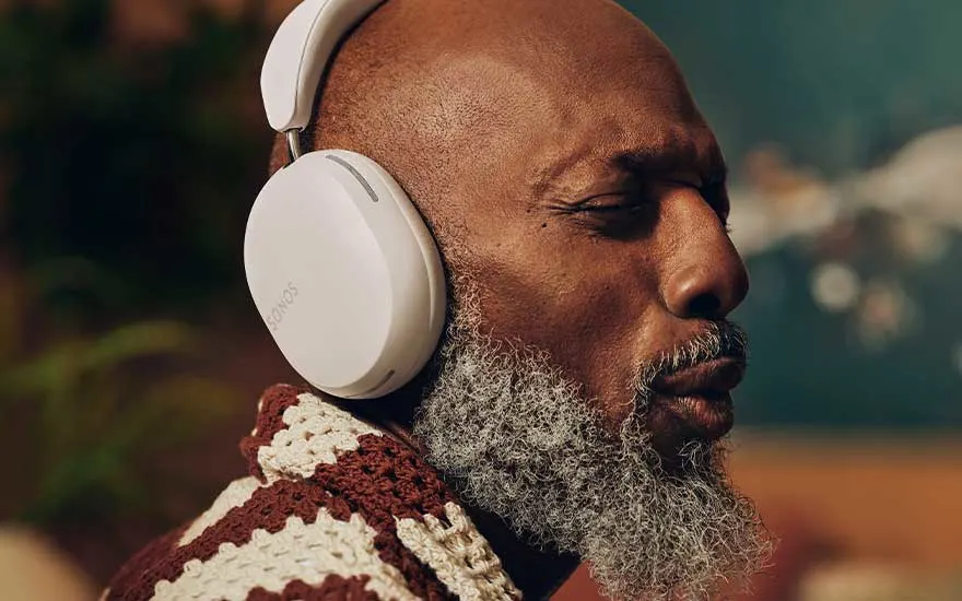 Een close-up van een witte Sonos hoofdtelefoon, gedragen met een bruin en wit gehaakt kledingstuk. De achtergrond is onscherp en kleurrijk.