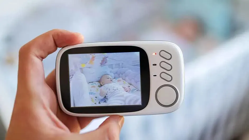 Main tenant un babyphone blanc avec écran LCD. L'écran montre un bébé couché dans un berceau.