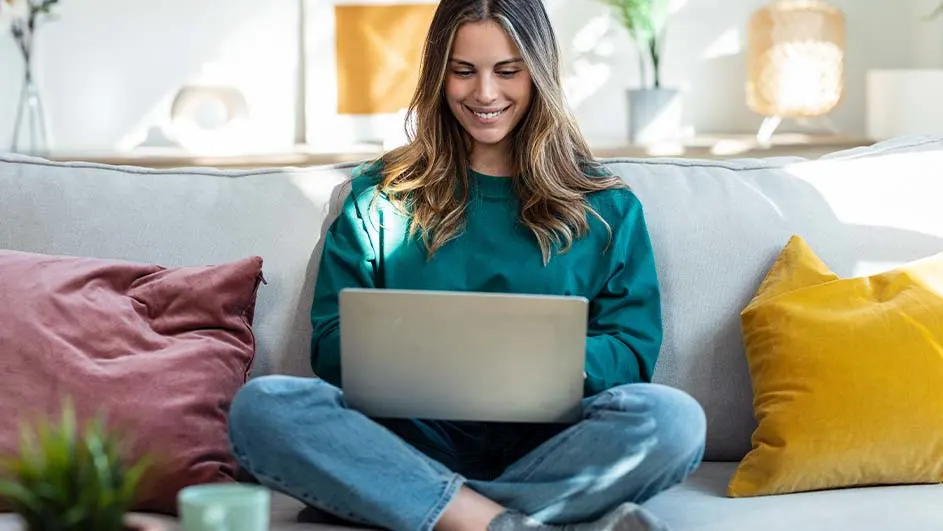 Een laptop op de schoot op een grijze sofa met roze en gele kussens. Planten en een lamp op de achtergrond.