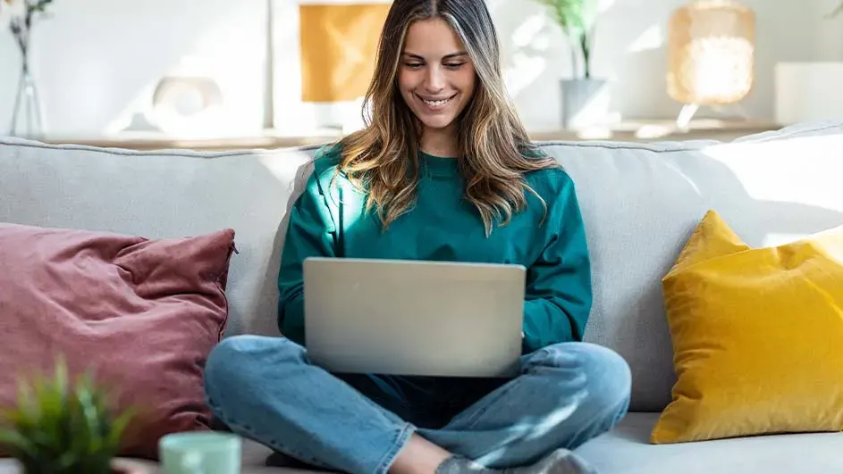 Een laptop op de schoot op een grijze sofa met roze en gele kussens. Planten en een lamp op de achtergrond.