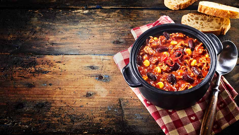 Een zwarte pot met chili staat op een rood-wit geruite doek, naast een zilveren lepel. Brood ligt op een rustieke houten tafel.