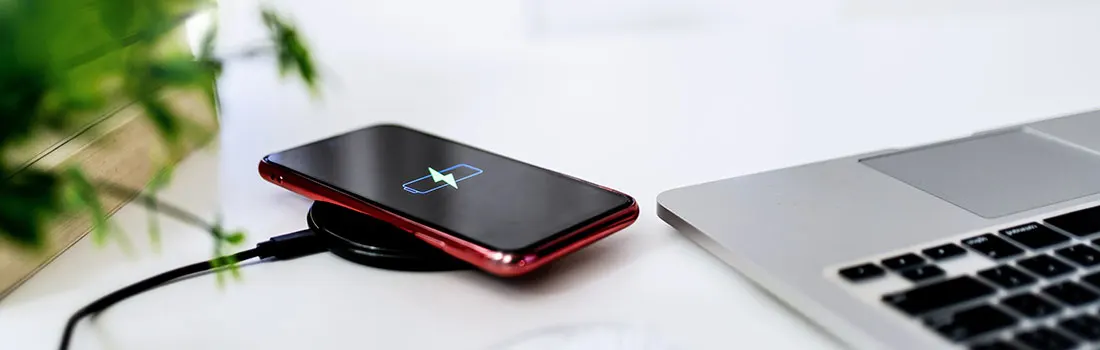 Un smartphone rouge charge sans fil sur un bureau blanc près d'un ordinateur portable et d'une plante.