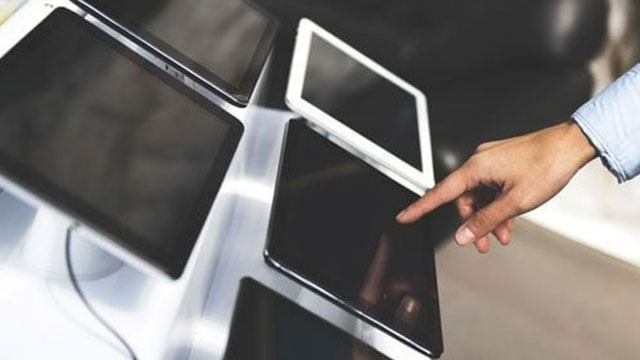 Plusieurs tablettes sur un présentoir argenté. Une main touche l'écran d'une tablette noire.