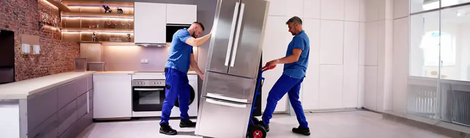 Deux déménageurs en bleu transportent un réfrigérateur en acier inoxydable dans une cuisine.