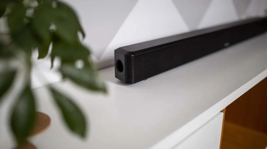 Zwarte soundbar op een witte plank tegen een witte muur met grijs geometrisch patroon. Groene plant links op de voorgrond.
