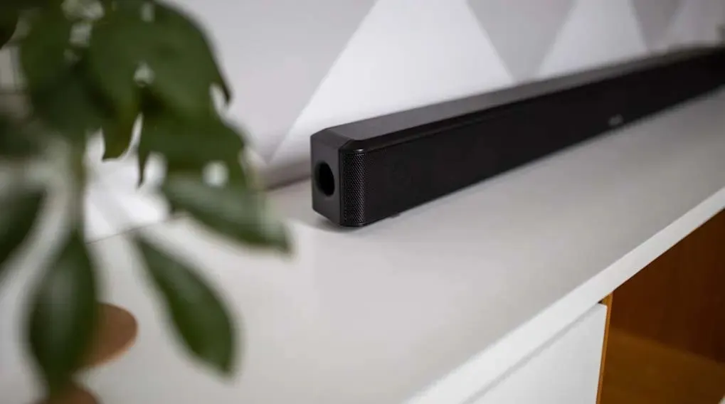 Zwarte soundbar op een witte plank tegen een witte muur met grijs geometrisch patroon. Groene plant links op de voorgrond.