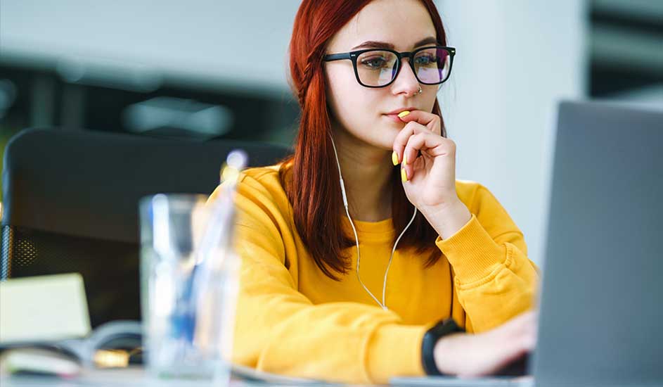 Une personne travaille sur un ordinateur portable gris, portant un pull jaune et des écouteurs blancs, sur un bureau blanc.