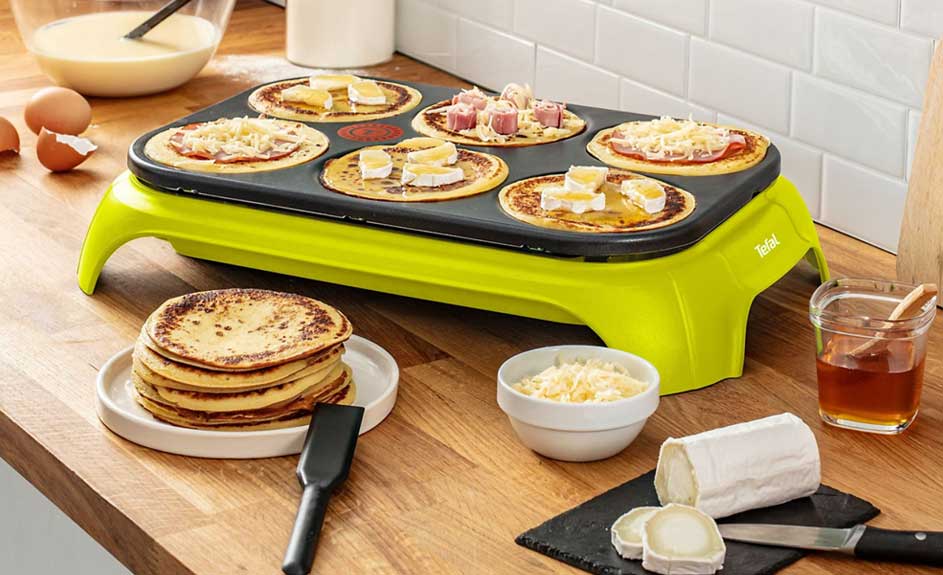 Gaufrier vert avec crêpes garnies sur une table en bois. Fromage, miel et bol de fromage râpé à côté.