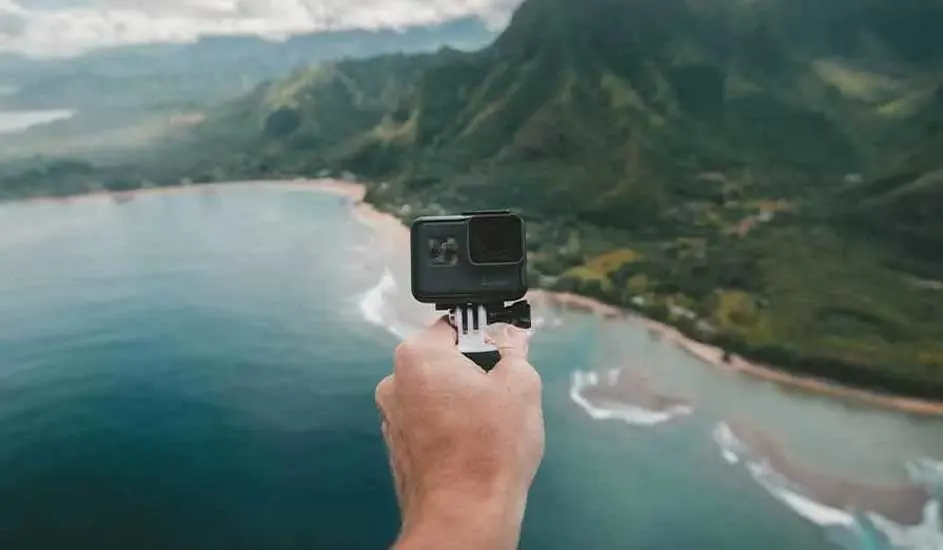 Een zwarte GoPro-camera wordt vastgehouden met een oceaan en groene bergen op de achtergrond.