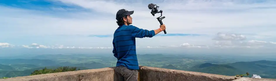 Un homme filme un paysage avec une caméra stabilisée. Le ciel est bleu avec des nuages.