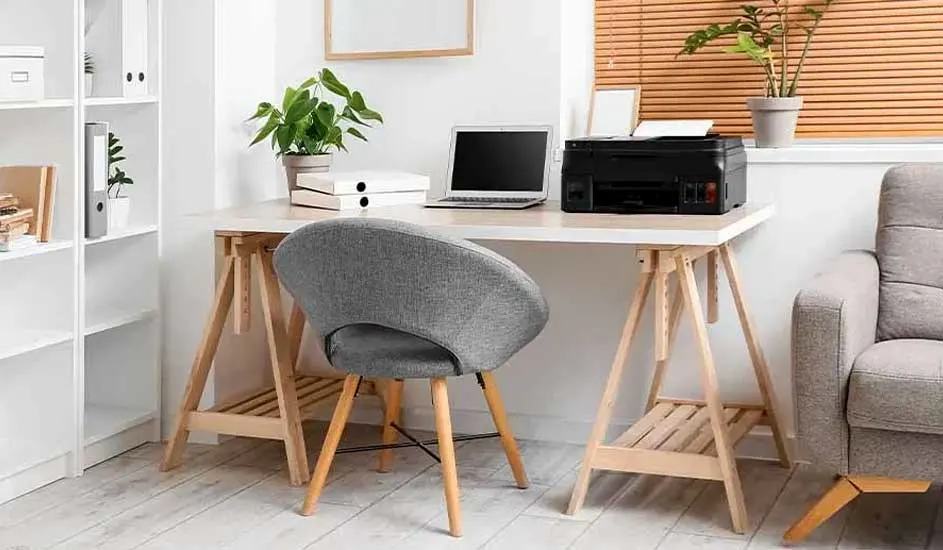 Bureau blanc avec ordinateur portable, imprimante, plante, chaise grise et étagère dans une pièce lumineuse.