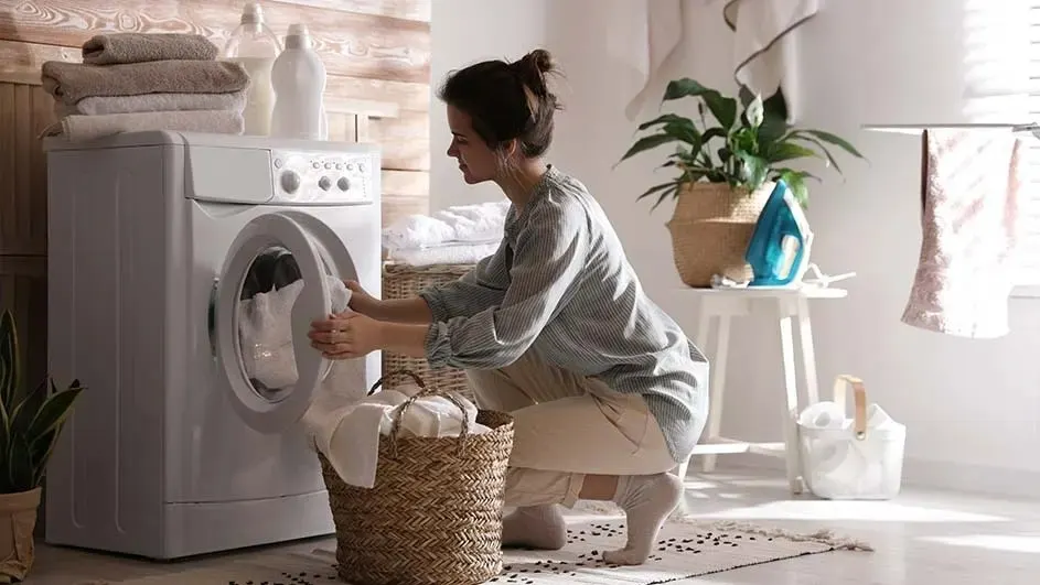 Een persoon plaatst witte handdoeken in een wasmachine in een lichte, moderne wasruimte met planten en een mand. Geen merk is zichtbaar.