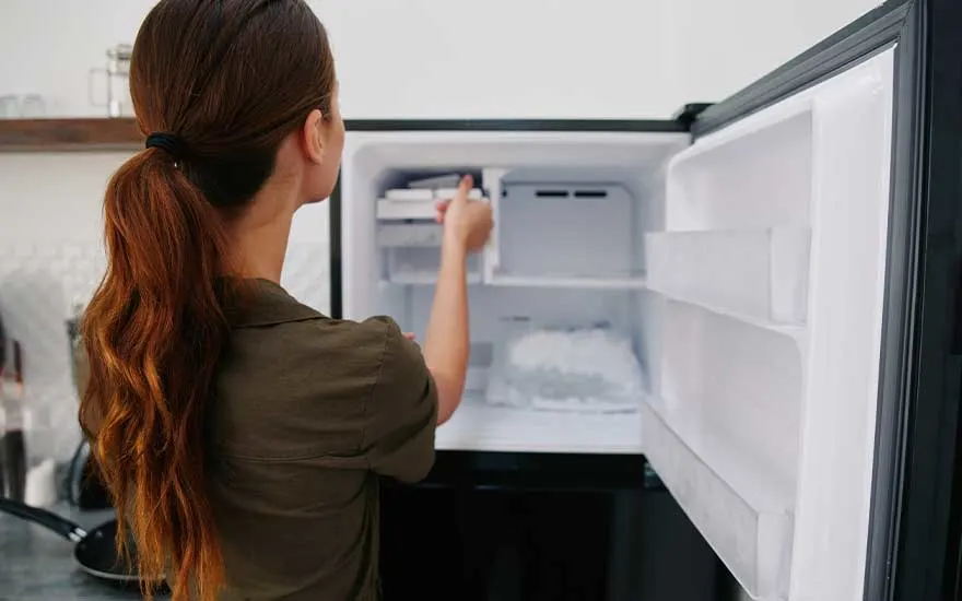 Een open koelkast met witte planken en een zwart frame, gevuld met ijs en bakjes. Iemand haalt iets uit de bovenste plank.