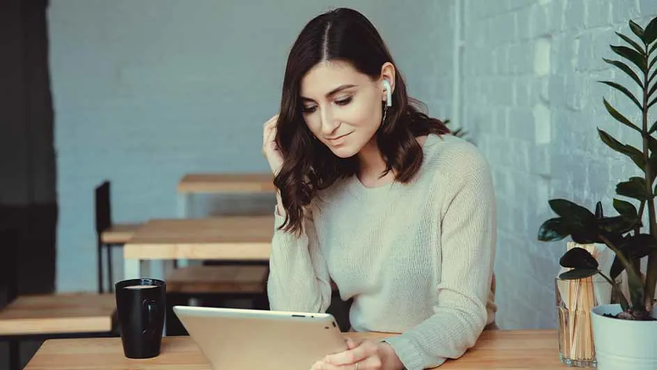 Een tablet op een houten tafel met een zwarte mok. Achtergrond: tafels, stoelen en een plant tegen een grijze muur.