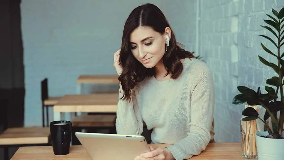 Une tasse noire, une tablette et une plante sont sur une table en bois. Des tables sont à l'arrière-plan, devant un mur bleu.