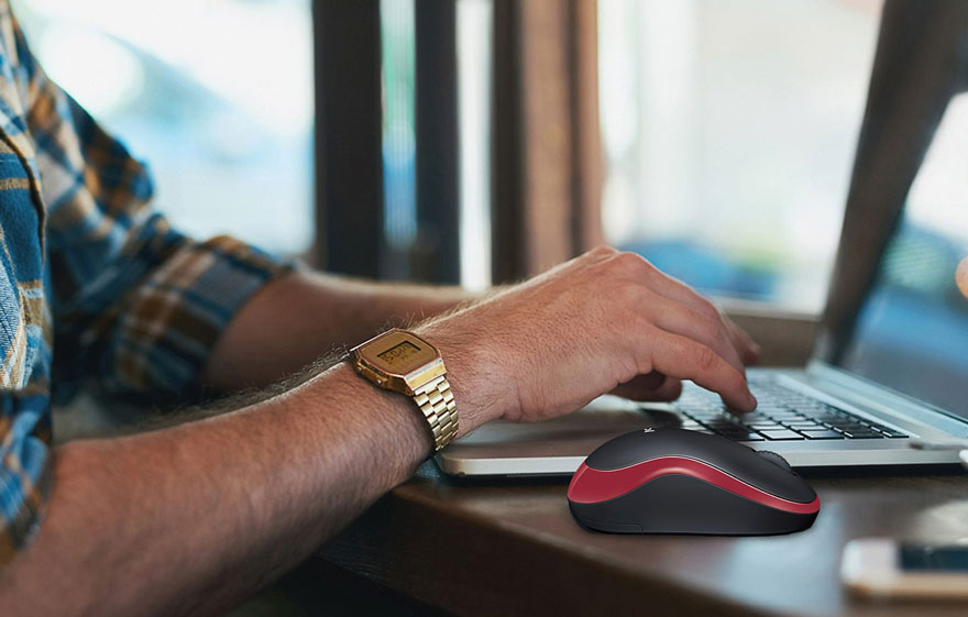 Une souris noire et rouge est posée à côté d'un ordinateur portable sur une table en bois, avec une montre dorée visible.