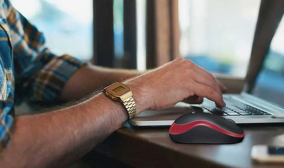 Persoon typt op een laptop naast een zwarte en rode draadloze muis. Een gouden horloge is zichtbaar.