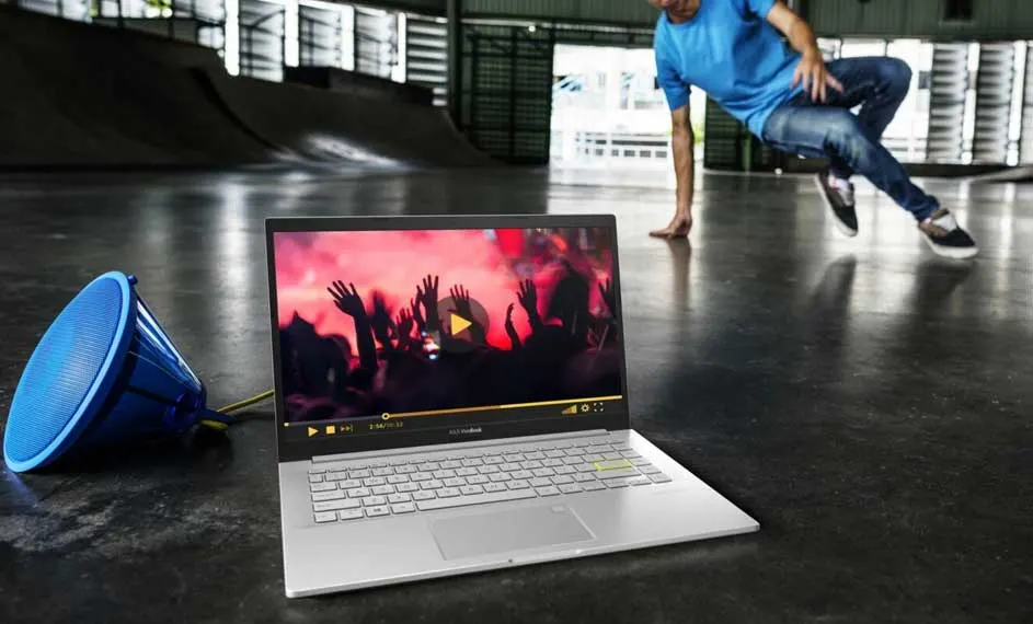 Een zilveren laptop speelt een video af, met een blauwe luidspreker ernaast, op een gepolijste betonnen vloer in een skatepark.