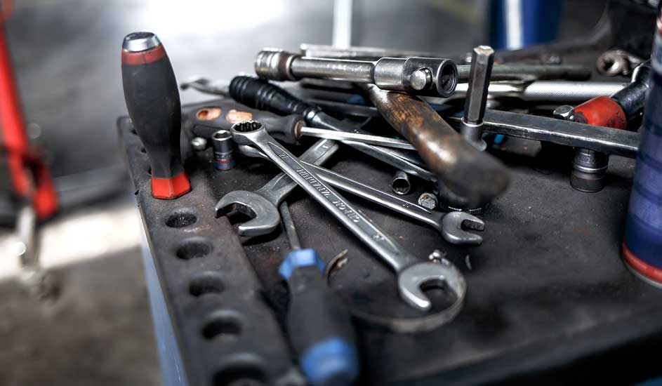 Een werkbank vol gereedschap. Sleutels, schroevendraaiers en een hamer liggen door elkaar op een donker oppervlak.