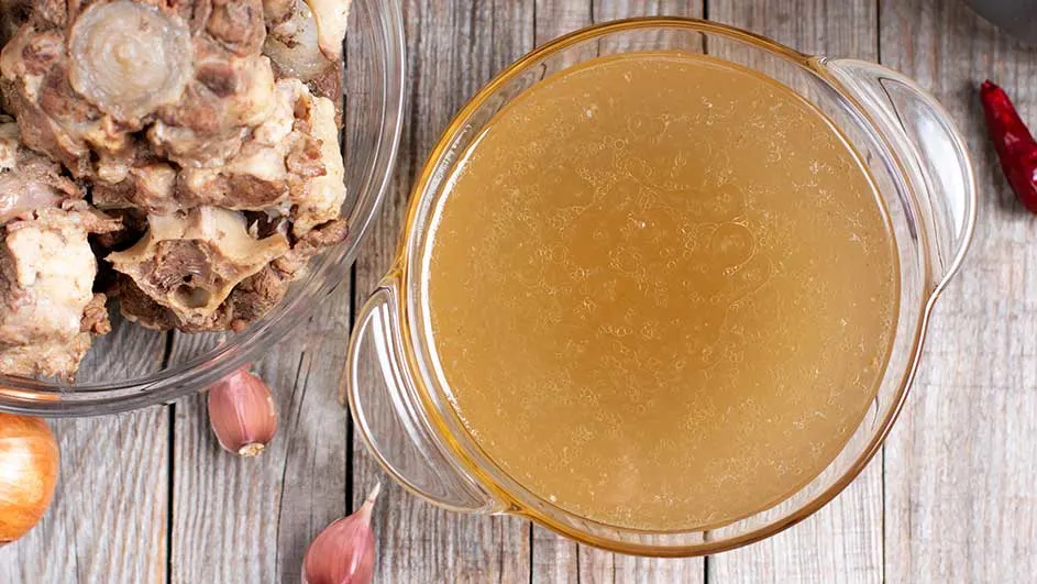 Een bovenaanzicht toont een glazen kom bouillon, een kom ossenstaart, knoflook en peper op een houten tafel.