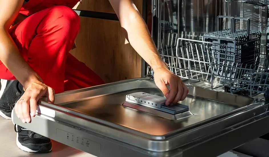 Een geopende vaatwasser met metalen rekken. Iemand in een rode broek controleert een grijs onderdeel in de vaatwasser.