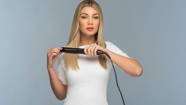 Une personne utilise un fer à lisser noir sur des cheveux blonds, sur un fond gris uni.