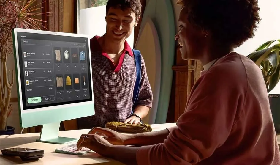 Two people at a counter use a mint Apple iMac as a shop till, browsing items and totals on screen, with a compact keyboard and mouse.
