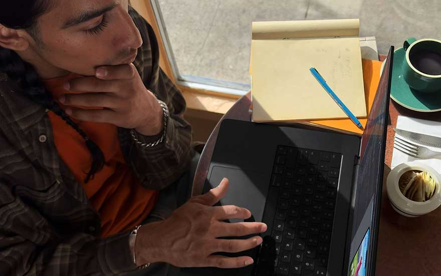 Een laptop staat op een tafel naast een notitieblok met een blauwe pen, een kopje koffie en bestek.