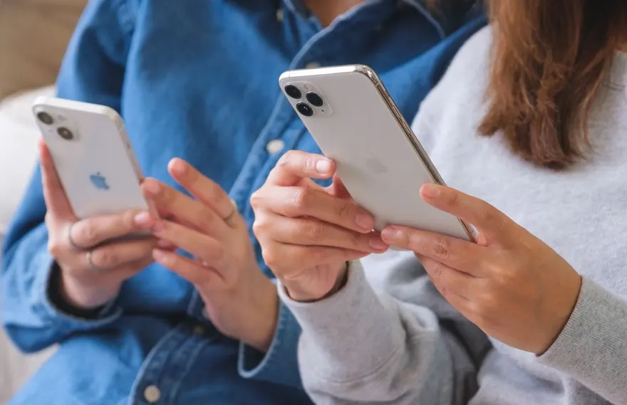Twee witte smartphones worden vastgehouden door handen tegen een achtergrond van blauwe en grijze kleding.