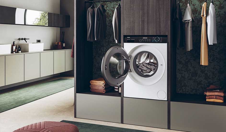 Une buanderie présente une machine à laver blanche ouverte dans un placard gris, avec des vêtements suspendus et des serviettes pliées.